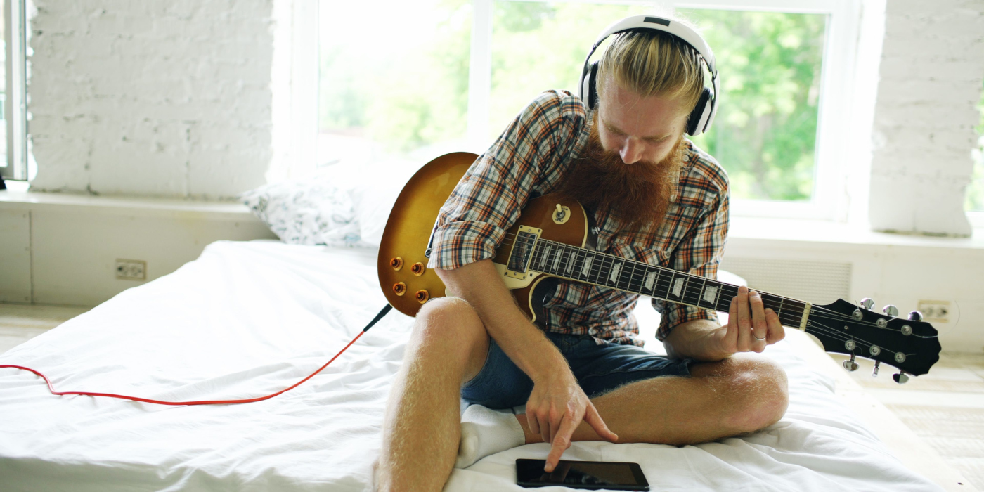 A middle aged man playing a Les Paul style guitar on a bed wearing headphones and scrolling through his phone