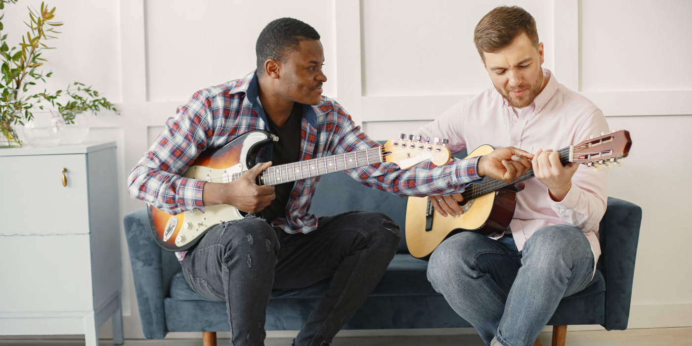 A man with an electric guitar teaching another man how to play a classical guitar while sitting on a sofa