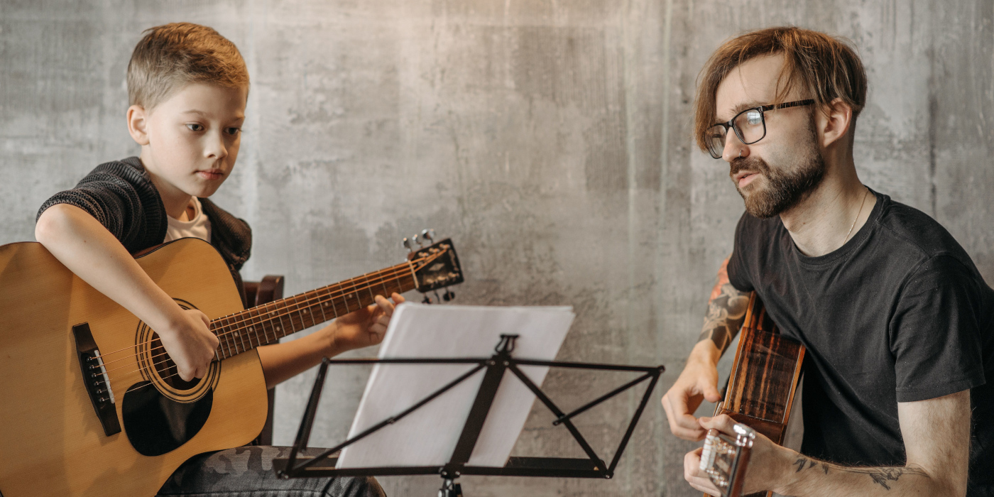 A young boy and male adult teacher playing acoustic guitars while reading sheet music on a music stand