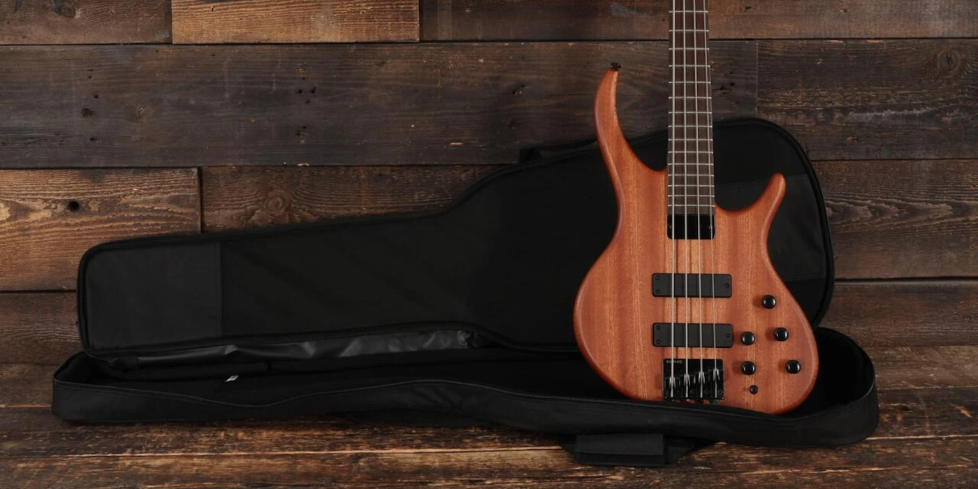 A Tobias Killer B IV bass guitar in a natural finish leaning up against a wooden wall sat in a black gig bag