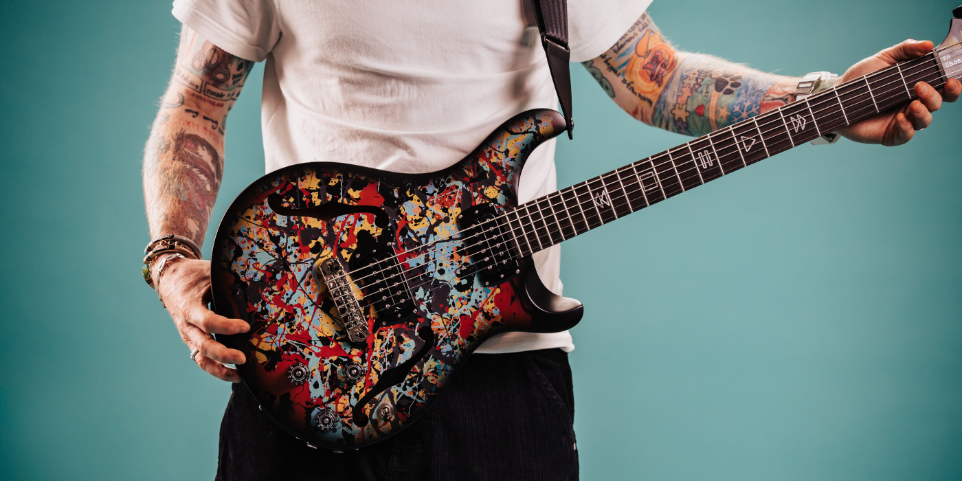 A studio blue background photo showing Ed Sheeran holding his PRS SE Cosmic Splash Limited Edition baritone electric guitar