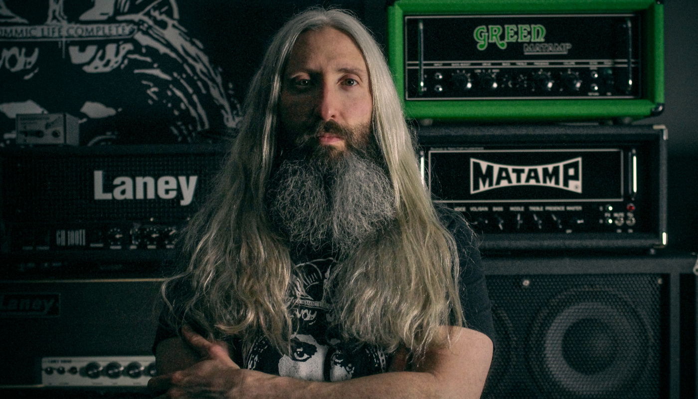 A photo of Does It Doom founder Steve Reis standing in front of several guitar amps with his arms folded