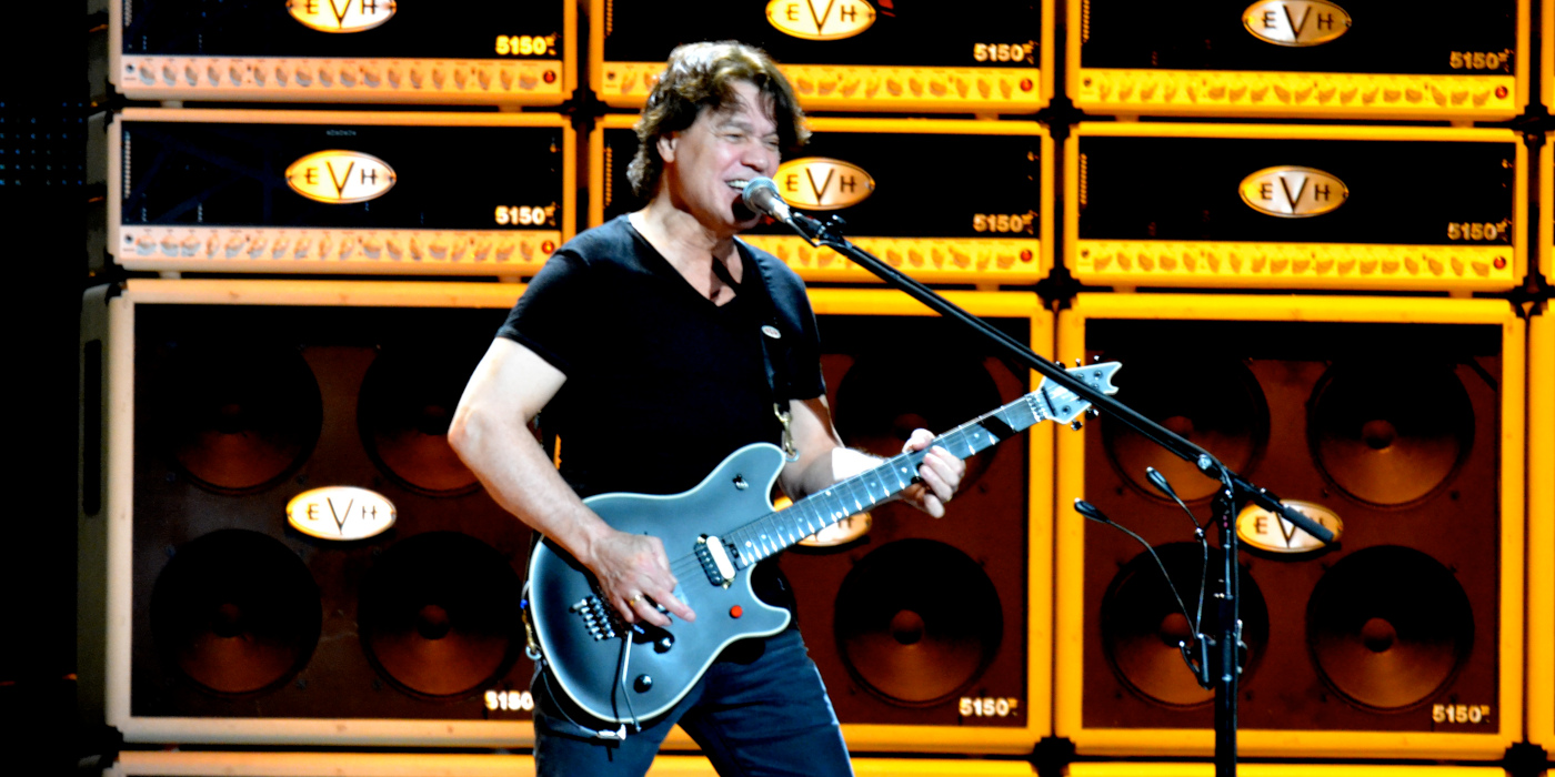 A photo of Eddie Van Halen on stage playing a black EVH Wolfgang Stealth guitar in front of a wall of EVH 5150 amps and cabs