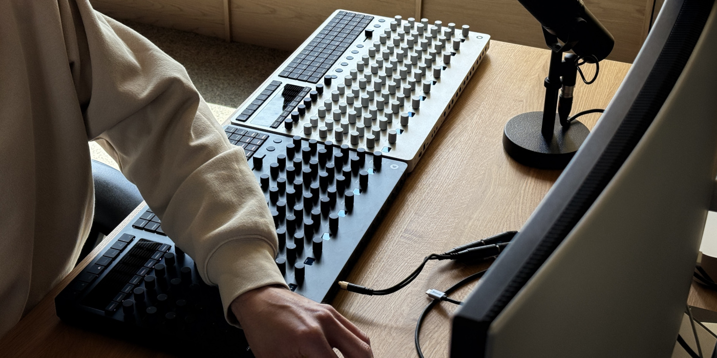 A lifestyle photo showing two Polyend Drums hybrid drum machines in black and silver on a desk in front of a computer monitor with a male working