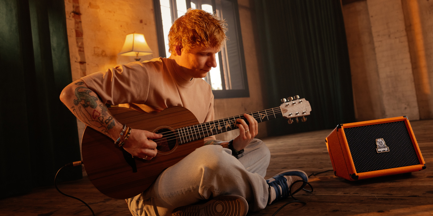 A lifestyle photo showing Ed Sheeran sat cross legged playing an acoustic guitar next to his signature Orange Outlowd ES60 amplifier