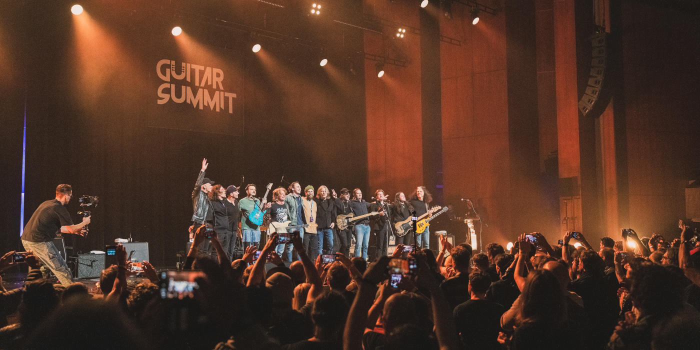 A photo of multiple famous guitarists standing on the stage at Guitar Summit with an energetic crowd