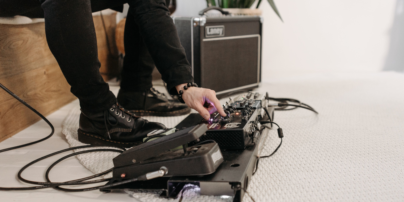 A pedalboard on a white rug in a lounge next to an amp with settings being changed by a person