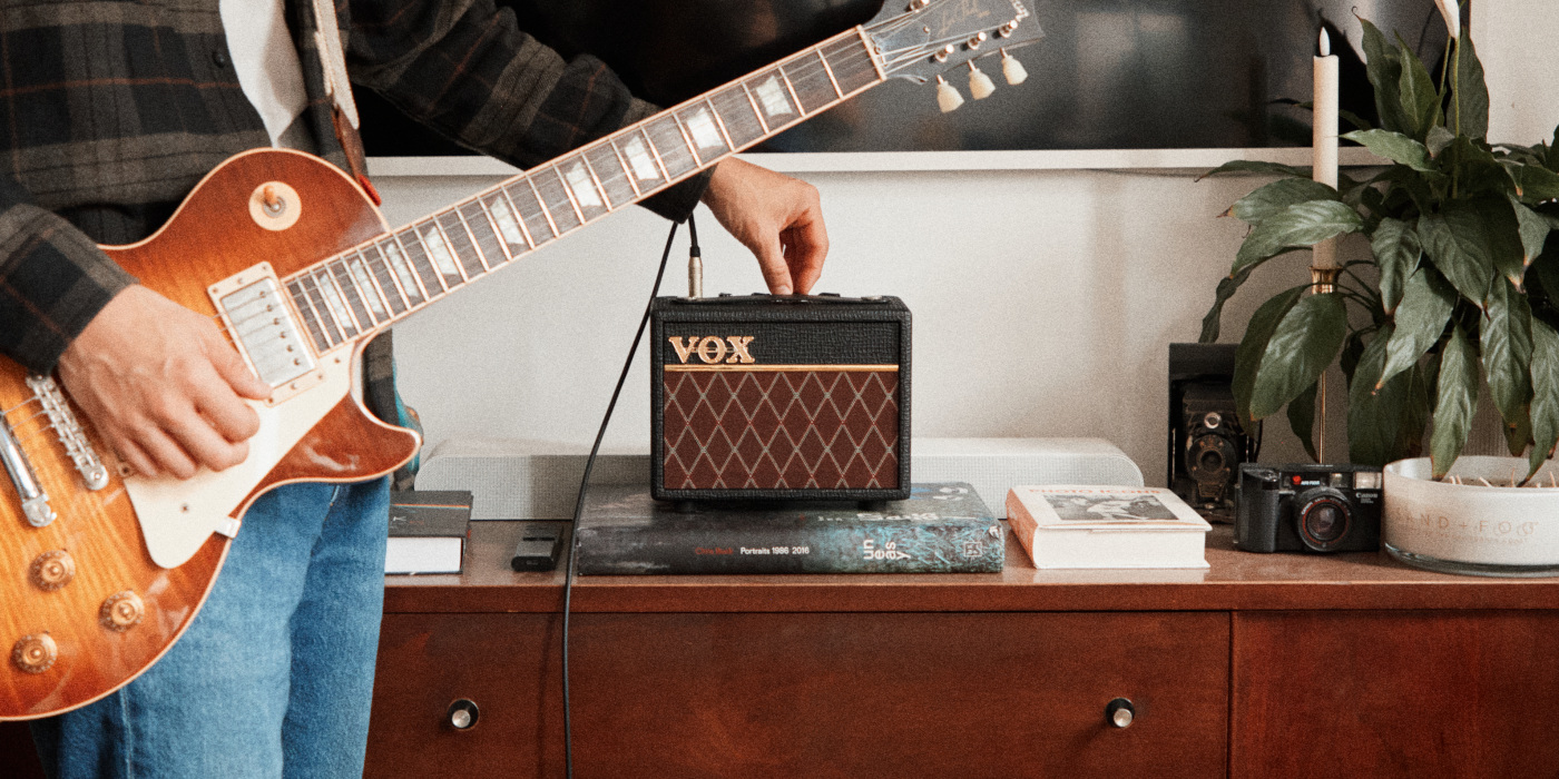 A guitarist adjusting the settings on a Vox AC Mini guitar amp that's placed on a sideboard while holding a Les Paul guitar