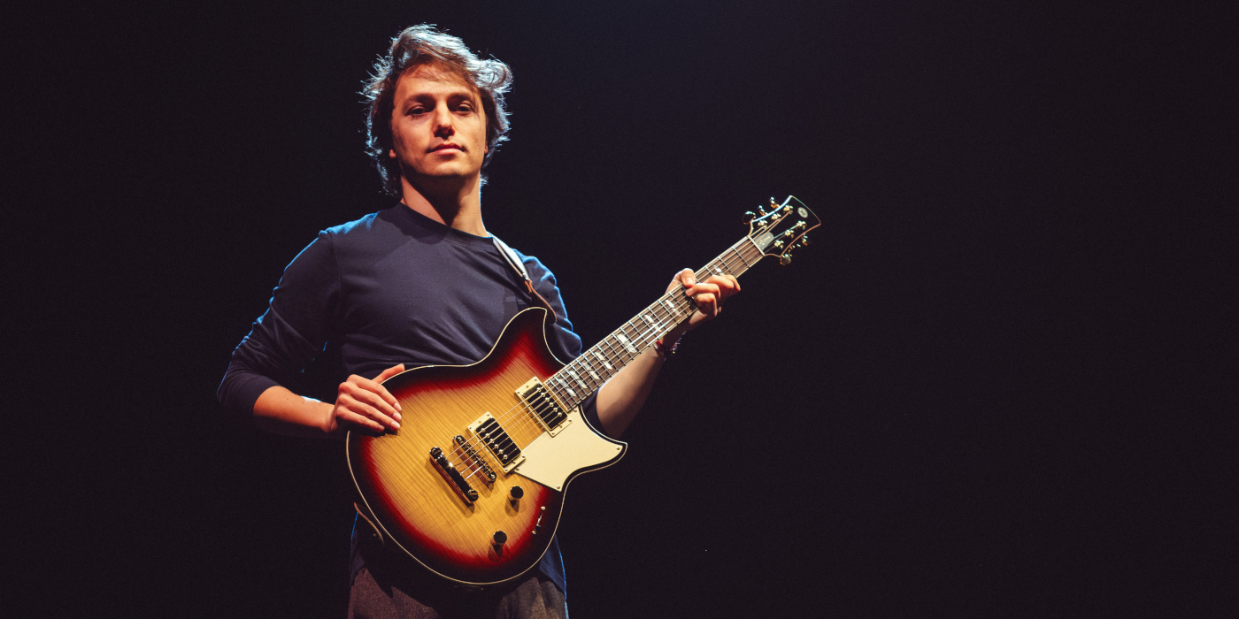 Matteo Mancuso holding his Yamaha Revstar RS20MM signature electric guitar with a dark background behind him