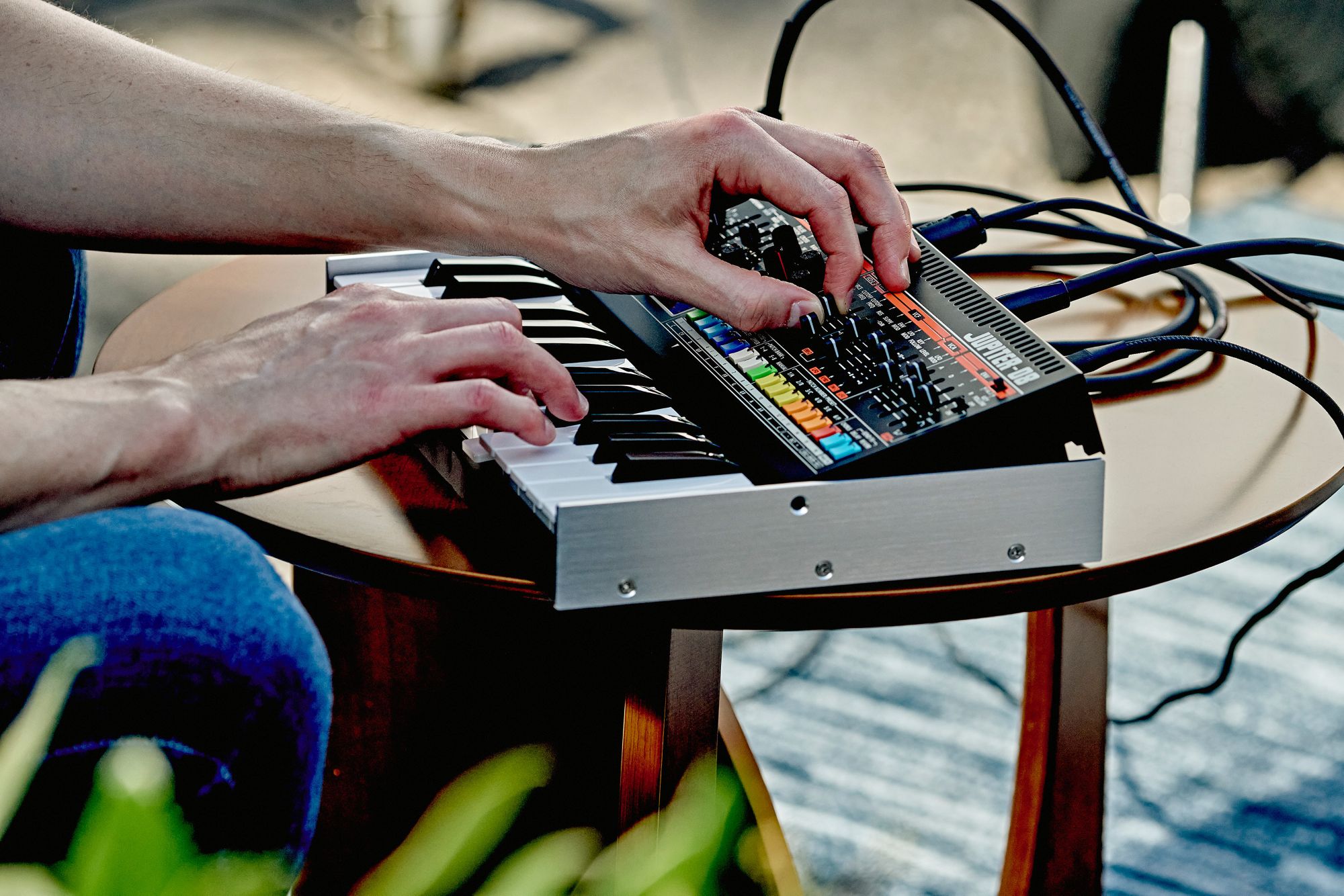 A picture of someone playing the Roland Jupiter-08 on a small table. It's connected up to various other pieces of equipment via MIDI, Audio and USB-C.