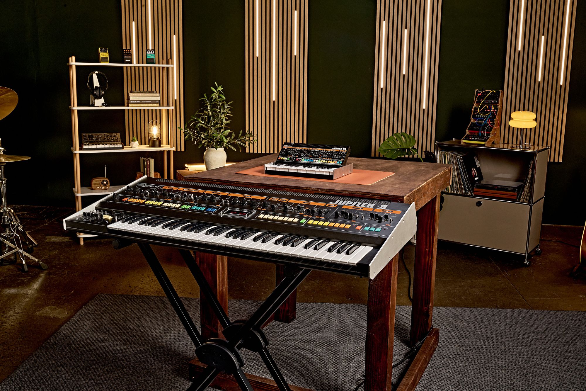 An original Roland Jupiter-8 synth in a music studio sat in front of the new Jupiter-08. In the background are are collection of Roland Micro Aira synths, Boss pedals, headphones and a Roland JP-08.